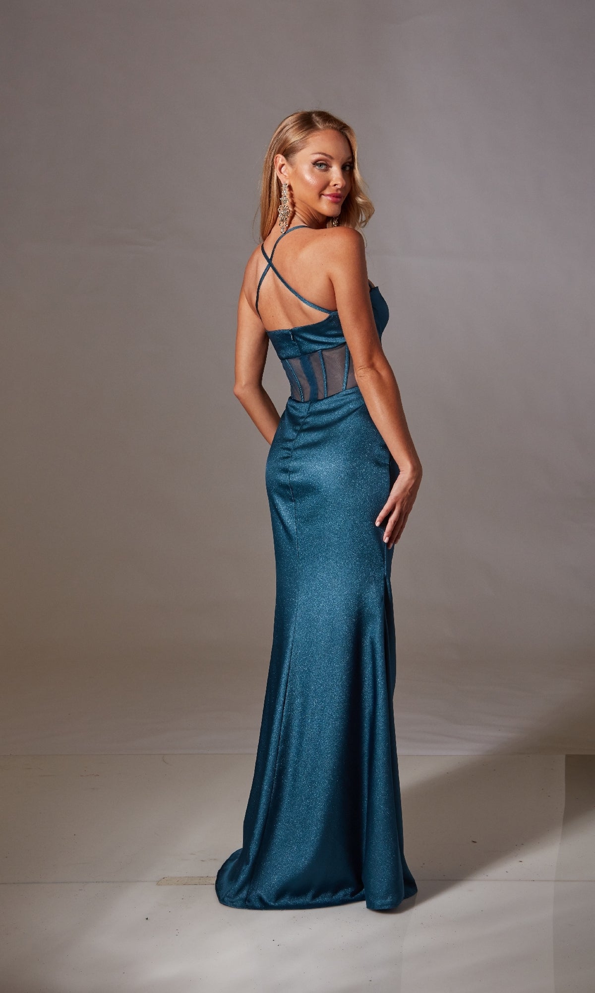 A woman in the Aerie Couture 250 Sheer-Corset Long Prom Dress with teal sheer paneling, a corset bodice, and crisscross straps stands backward, glancing over her shoulder against a plain background.
